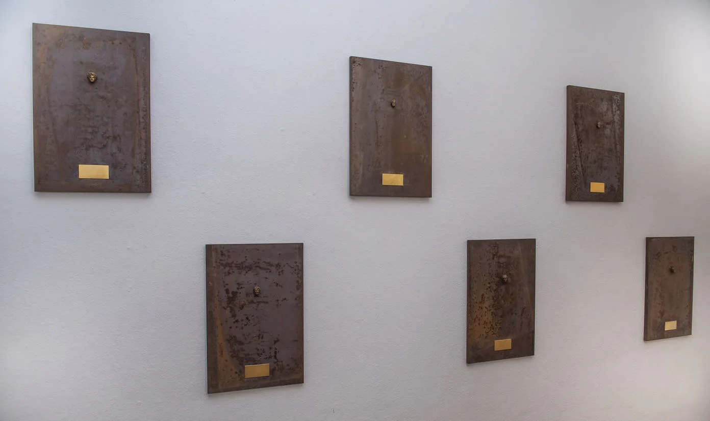 Cheats and Gods, Vernissage: sechs kleine rostige Metalltafeln mit Messingplaketten an einer weißen Wand