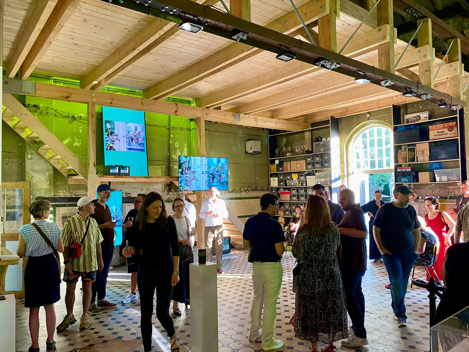 Neue Erinnerungen, Eröffnung: Besucher in der hellen Halle mit Holzdecke, Bildschirmen und einer Bücherwand