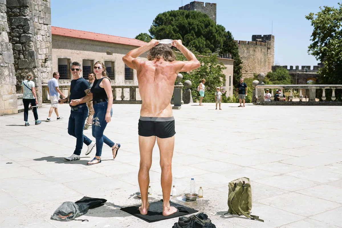 GABRIEL: Mann in Badehose steht mit nassem Rücken auf einem Platz vor einer Burgmauer, Kleider am Boden, Passanten schauen