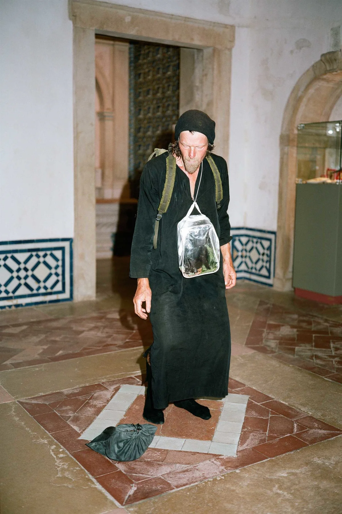 GABRIEL: Mann in Schwarz mit Beutelchen um den Hals steht in einem Raum mit Azulejos, Zweige am Boden
