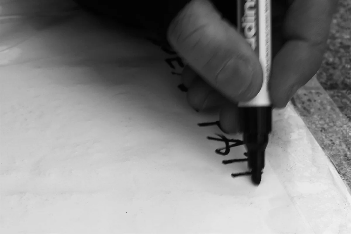 GABRIEL: Nahaufnahme einer Hand, die mit Filzstift Buchstaben auf Stein schreibt, Schwarz-Weiß