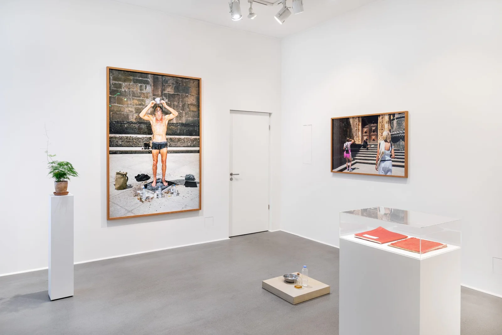 Installationsansicht: weißer Raum mit der Fotografie eines Mannes in Badehose, einer weiteren gerahmten Fotografie und einer Vitrine mit roten Büchern