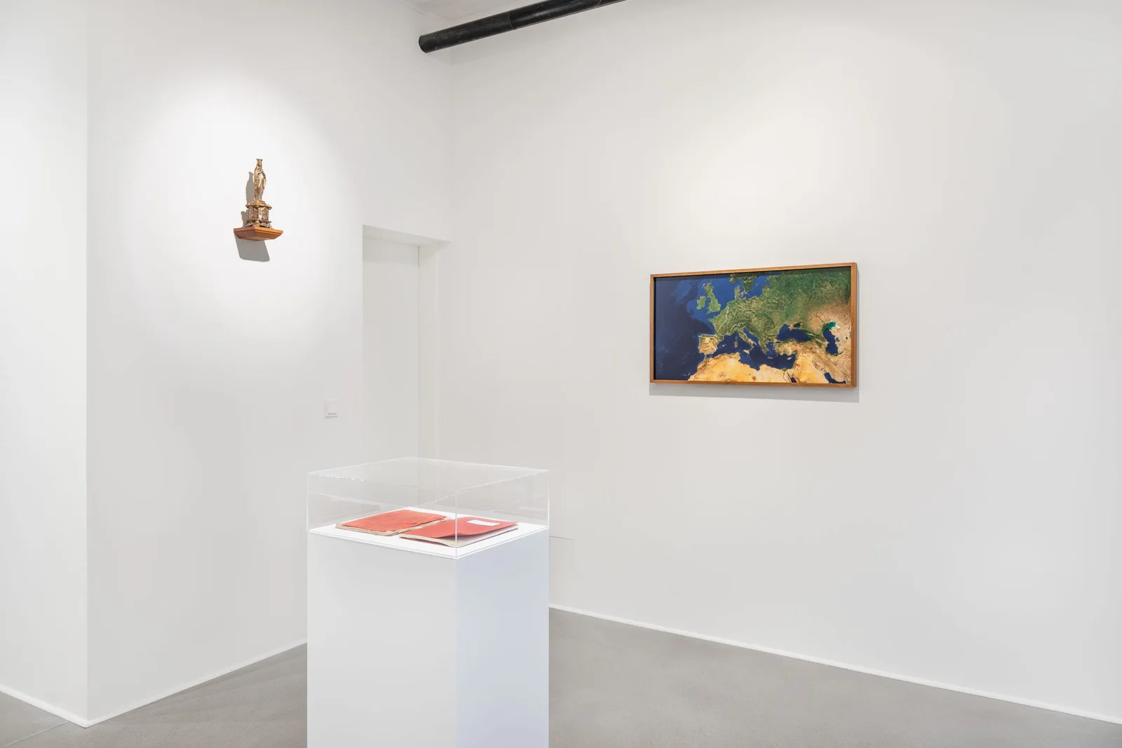 Installationsansicht: weißer Raum mit einer Madonnenfigur an der Wand, einer Satellitenkarte Europas und einer Vitrine mit roten Büchern