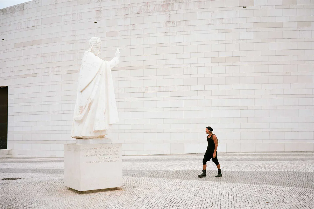 GABRIEL: Mann geht an einer weißen Statue vor der hellen Wand einer modernen Kirche vorbei