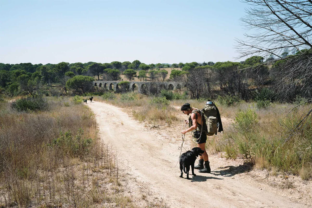 GABRIEL: Wanderer mit Rucksack und Hunden auf einem sandigen Weg, dahinter ein Aquädukt