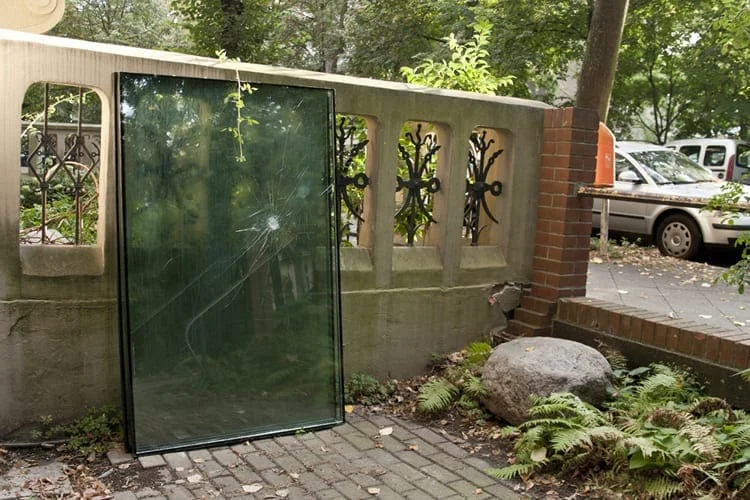 Große Glasscheibe, an eine Gartenmauer mit Gitterzaun gelehnt, dahinter eine Straße mit Autos