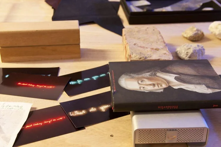 Tisch mit kleinem gerahmtem Porträt, schwarzen Tafeln mit roten und blauen Leuchtschriften, Steinen und Holzkästen