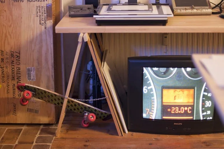 Ein Röhrenfernseher zeigt ein Armaturenbrett mit Tachometer und Temperaturanzeige, daneben ein Skateboard und eine Holzkiste