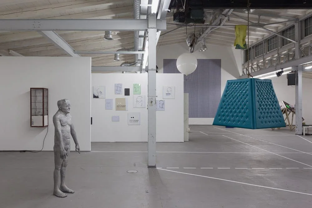 Installationsansicht: Industriehalle mit einer stehenden Figur, Bildern an einer Stellwand und einem türkisfarbenen Pyramidenobjekt