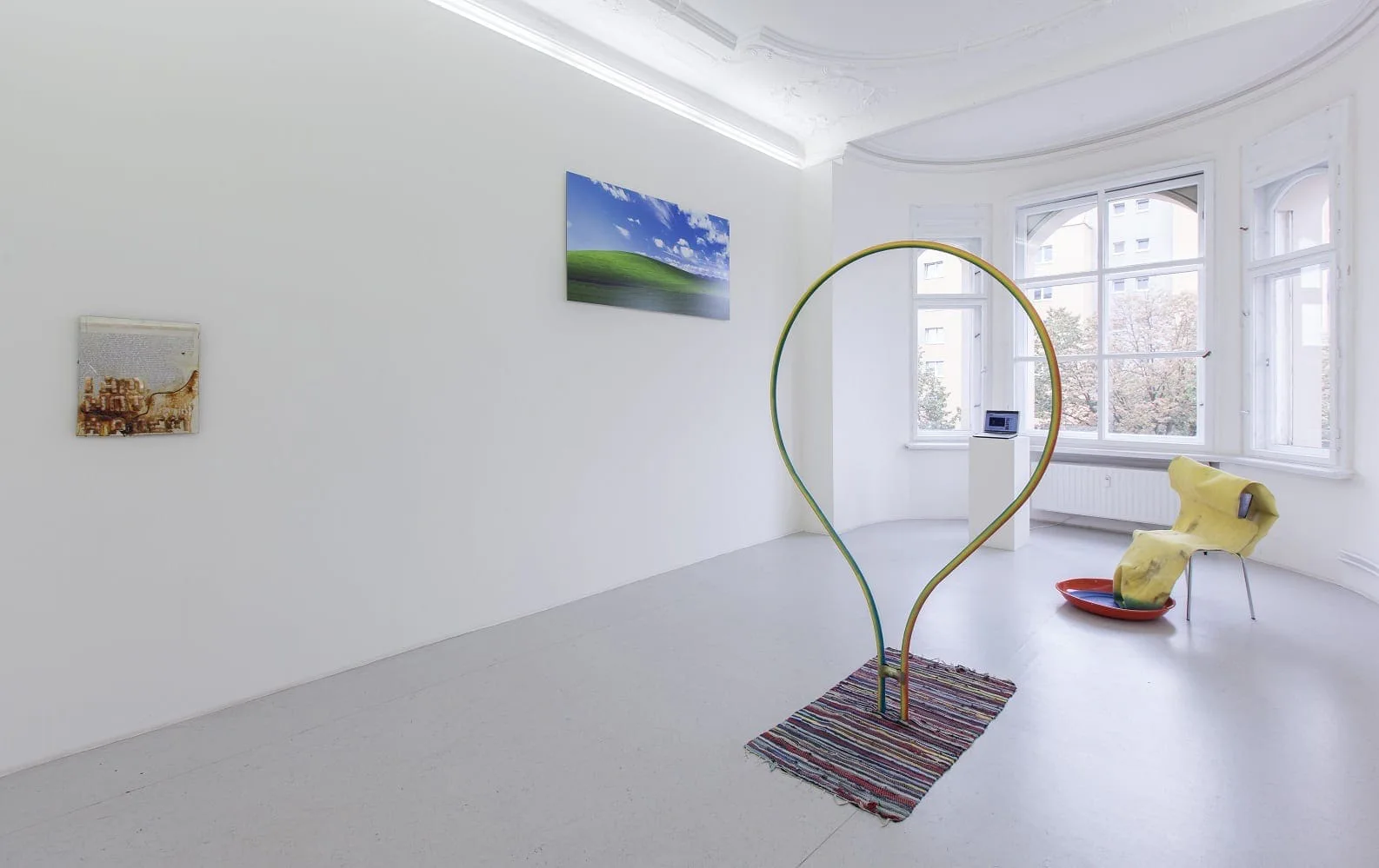 Installationsansicht mit Werken von Christoph Medicus, Mike Ruiz und Johannes Flechtenmacher: Bilder an der Wand, eine Schlaufe aus Holz auf einem Teppich, ein gelbes Objekt