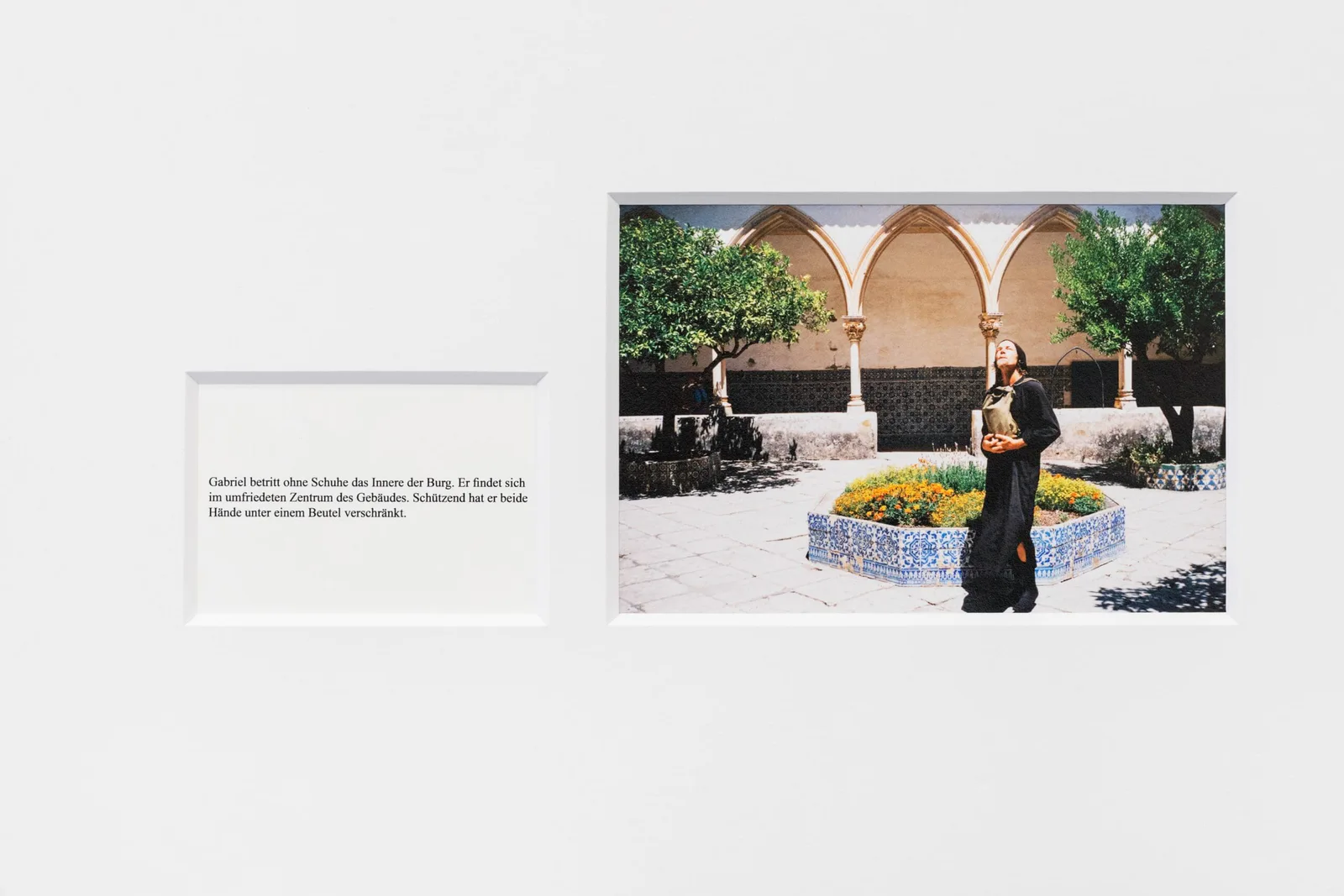 Marianische Antiphonen I, Ausschnitt: gerahmte Fotografie einer Frau in Schwarz in einem Arkadenhof mit Blumenbeet, daneben eine Texttafel