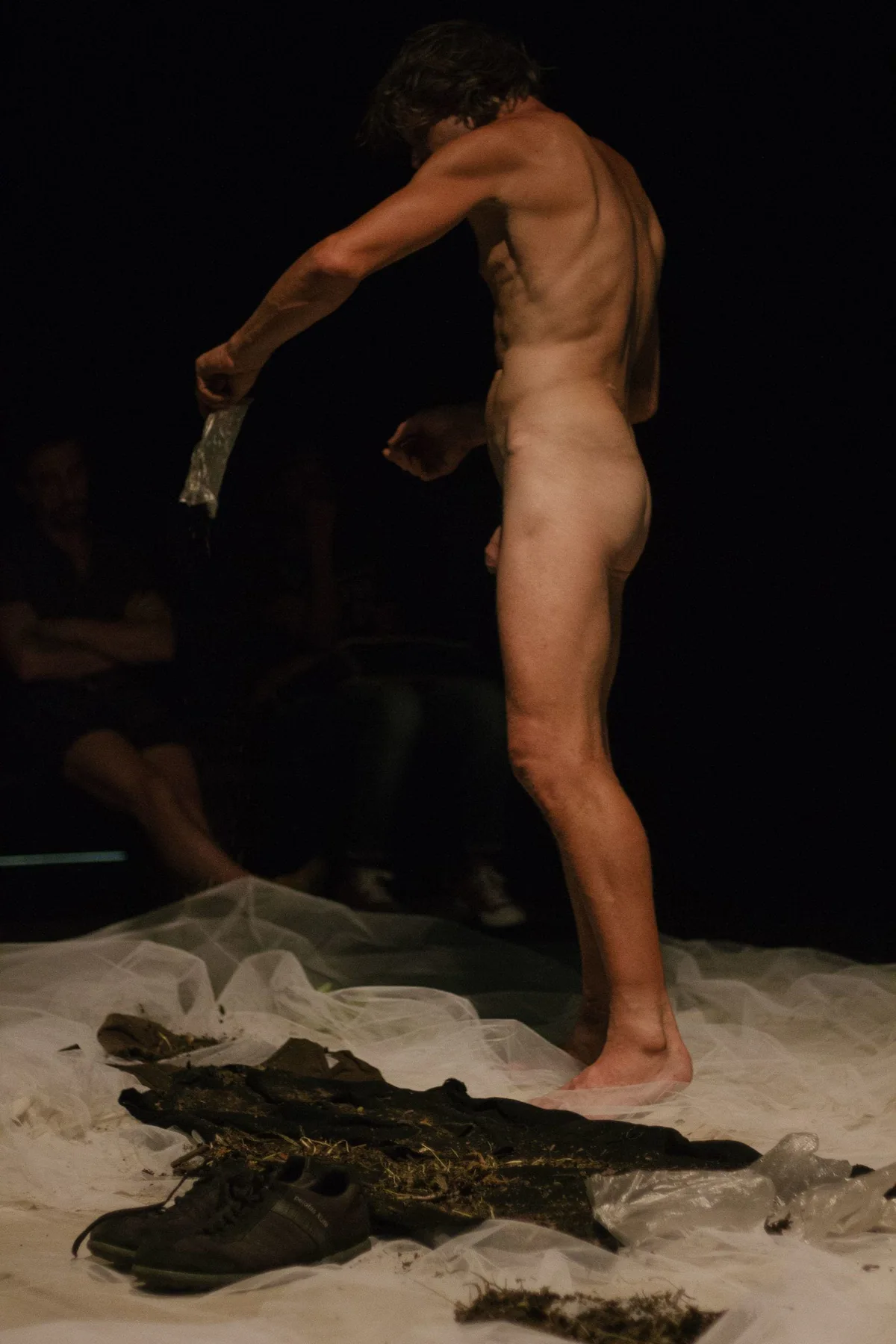 GABRIEL, Bühnenperformance: nackter Performer von hinten, Kleidung und Erde am Boden