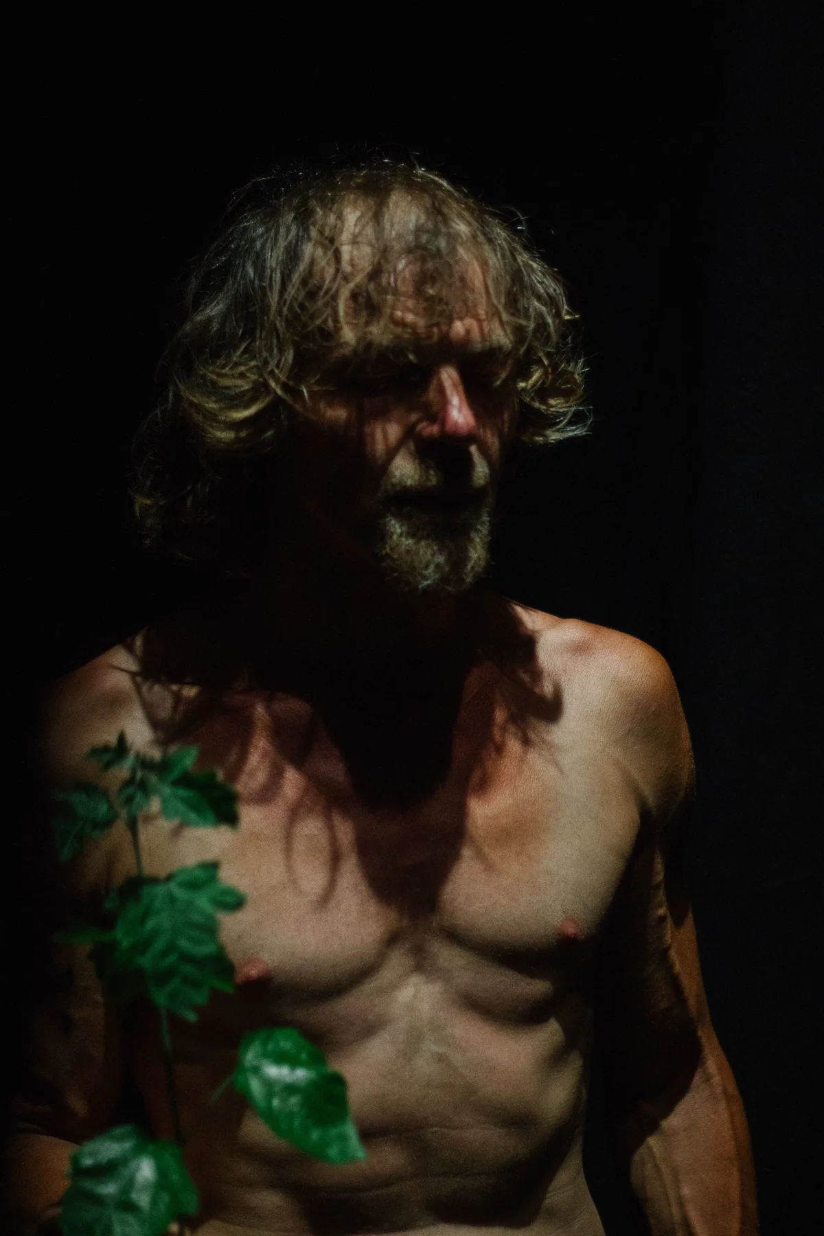 GABRIEL, Bühnenperformance: Porträt des Performers mit Blättern auf der nackten Brust im Dunkeln