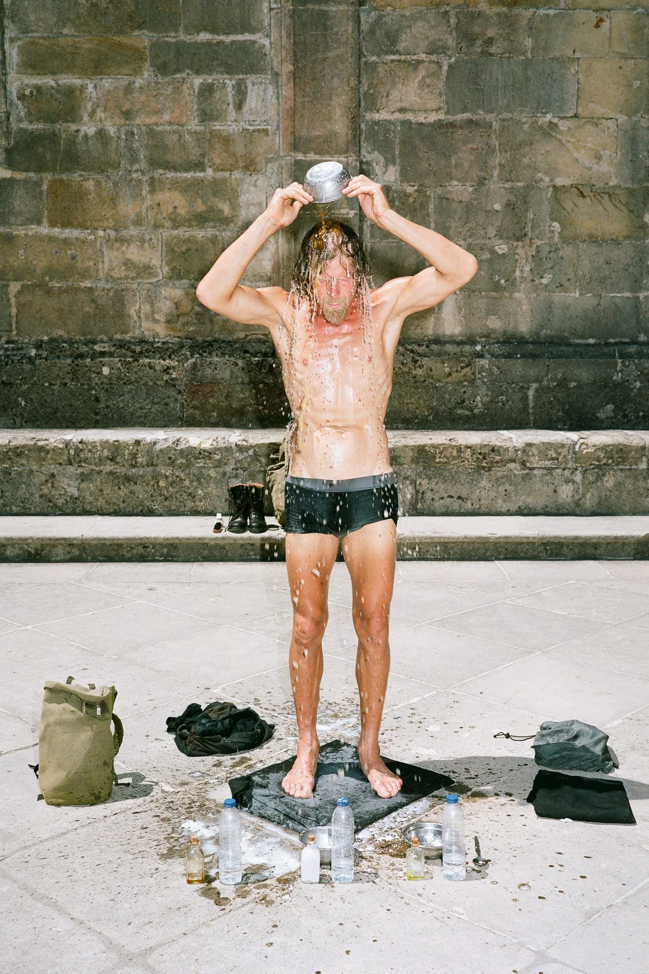 Gabriel & Stefan Hähnel: M.A. I – Die Dritte Waschung oder die Honigtaufe: ein Mann in Badehose gießt sich vor einer Steinmauer Wasser über den Kopf