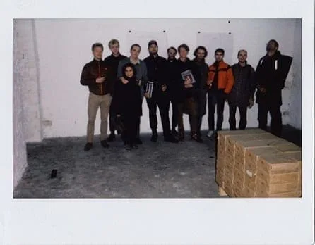 Max Schaffer – In Erwartung des Kommenden Gottes: Polaroid einer Gruppe von Menschen hinter gestapelten Holzkisten in einem Galerieraum