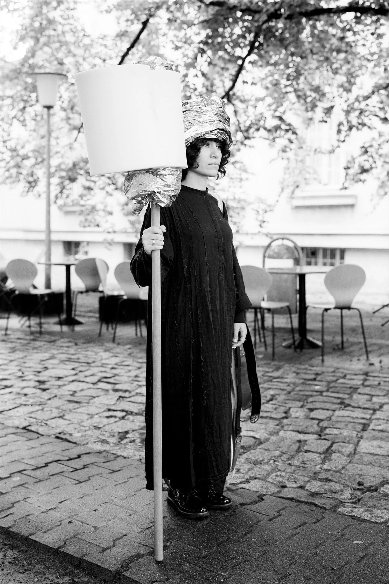 MNEMOSYNE Prelude – Die Könige – Letter of Politics or Rules for Kings: Performerin in schwarzem Gewand mit Stange und Lampenschirm in einem Innenhof, Schwarz-Weiß