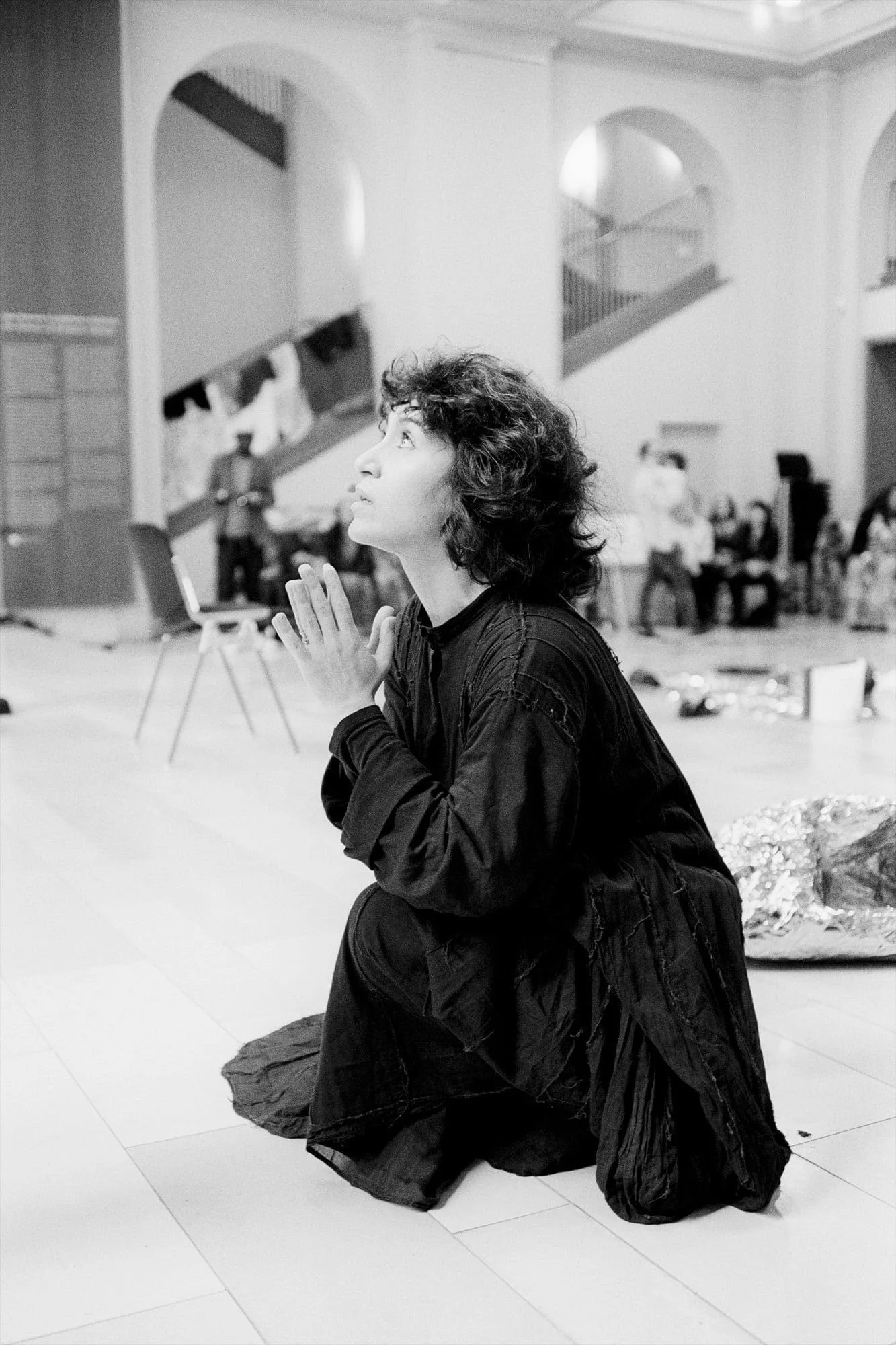 MNEMOSYNE Prelude – Die Könige – Letter of Politics or Rules for Kings: Performerin kniet in einem Saal mit Oberlichtern, Publikum im Hintergrund, Schwarz-Weiß