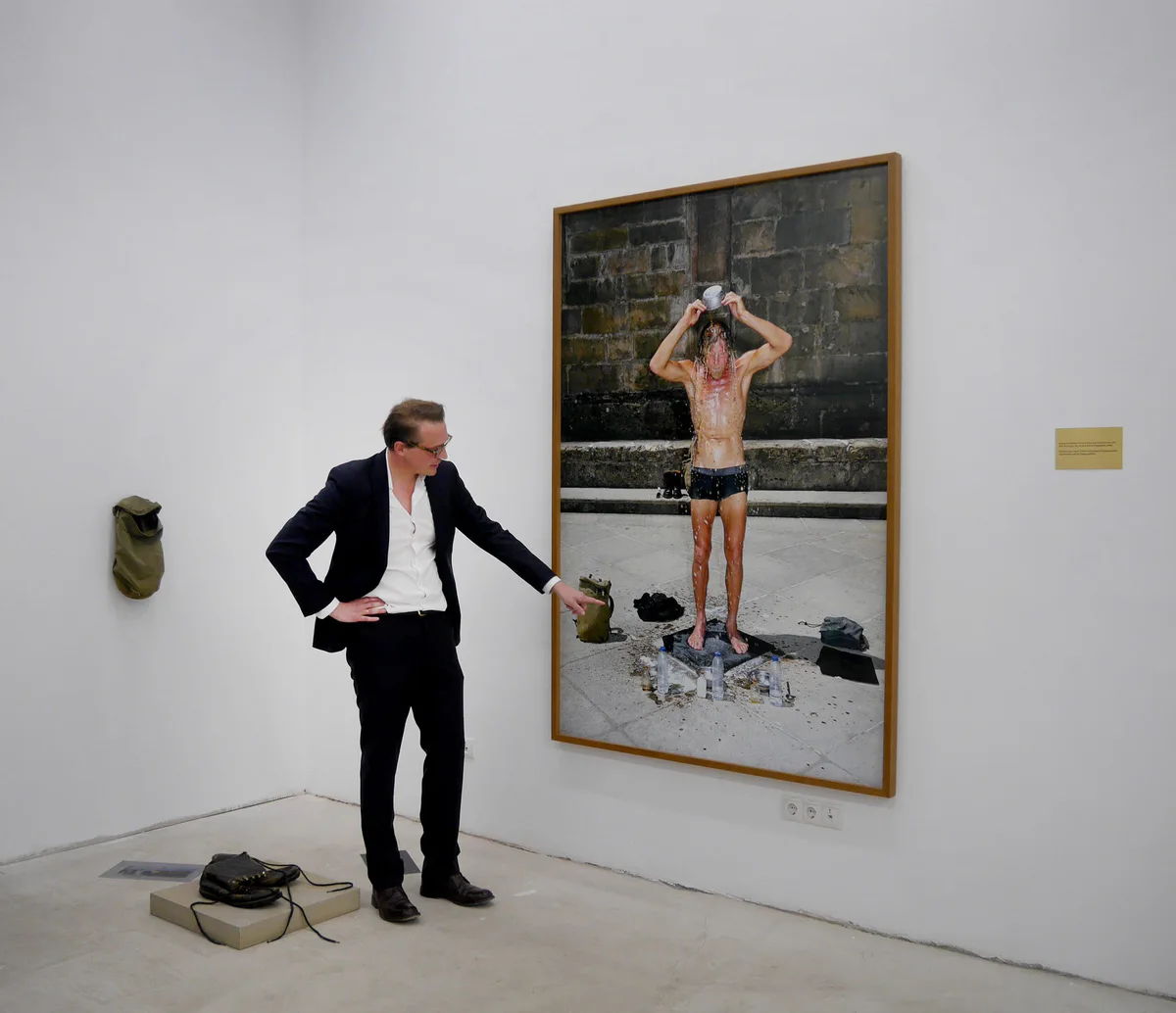 GABRIEL, Ausstellung: Mann im Anzug zeigt auf eine große Fotografie eines Mannes, der sich Wasser über den Kopf gießt
