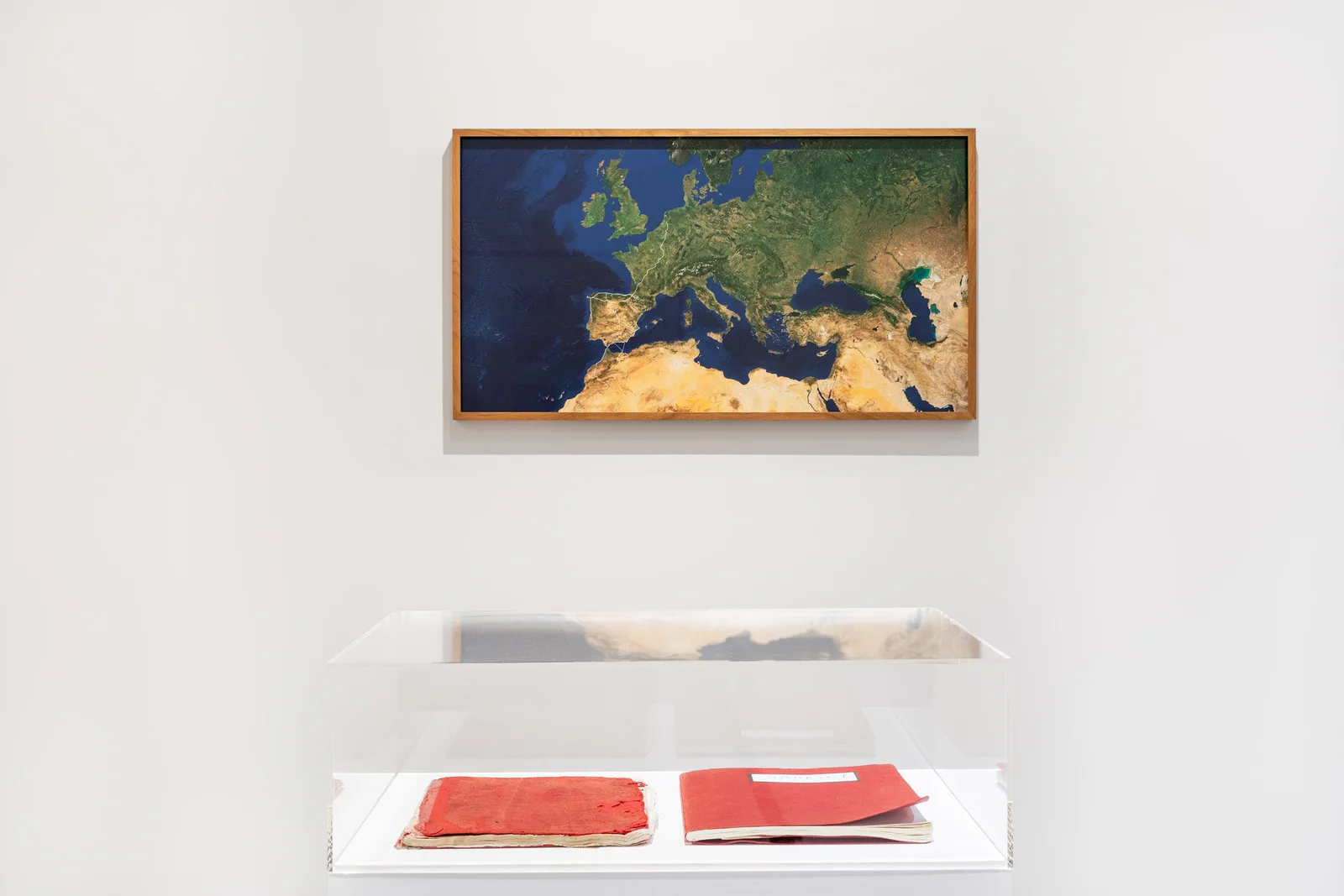 Mυθοποίηση – Mythopoesis: eine gerahmte Satellitenkarte Europas an der Wand, darunter eine Vitrine mit roten Büchern