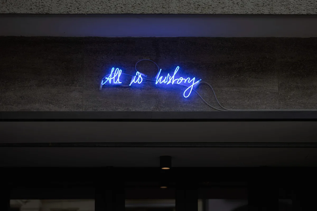 Tempel auf Zeit: blaue Neonschrift „All is history“ in einem dunklen Raum