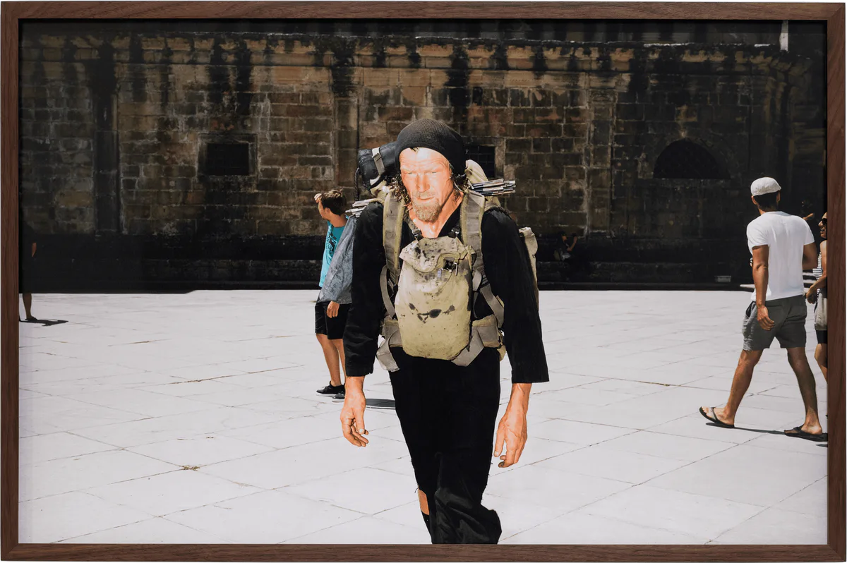 GABRIEL – Der Soldat der keinen Krieg führen wollte, 2019: gerahmte Fotografie eines Mannes mit Rucksack und Hut auf einem Platz vor einer Steinfassade