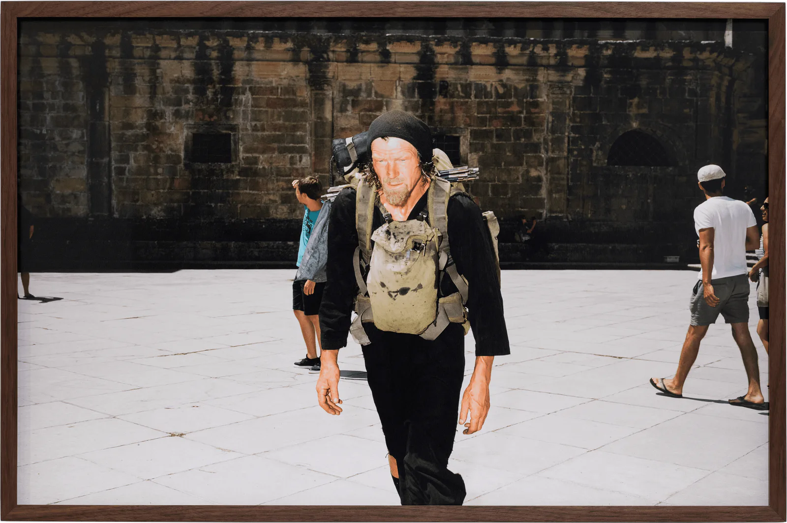GABRIEL – Der Soldat der keinen Krieg führen wollte, 2019: gerahmte Fotografie eines Mannes mit Rucksack und Hut auf einem Platz vor einer Steinfassade