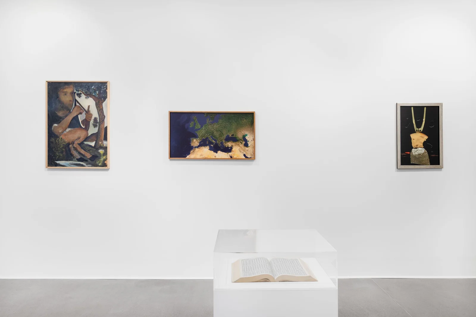 Ausstellungsansicht: drei Gemälde von Hilarion Manero an einer weißen Wand, davor ein aufgeschlagenes Buch auf einem Sockel
