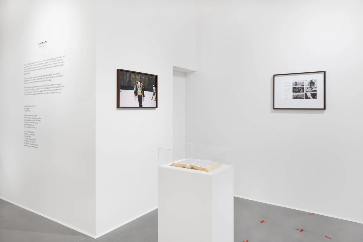 Ausstellungsansicht: weißer Raum mit Wandtext, zwei gerahmten Fotografien und einem flachen Sockel