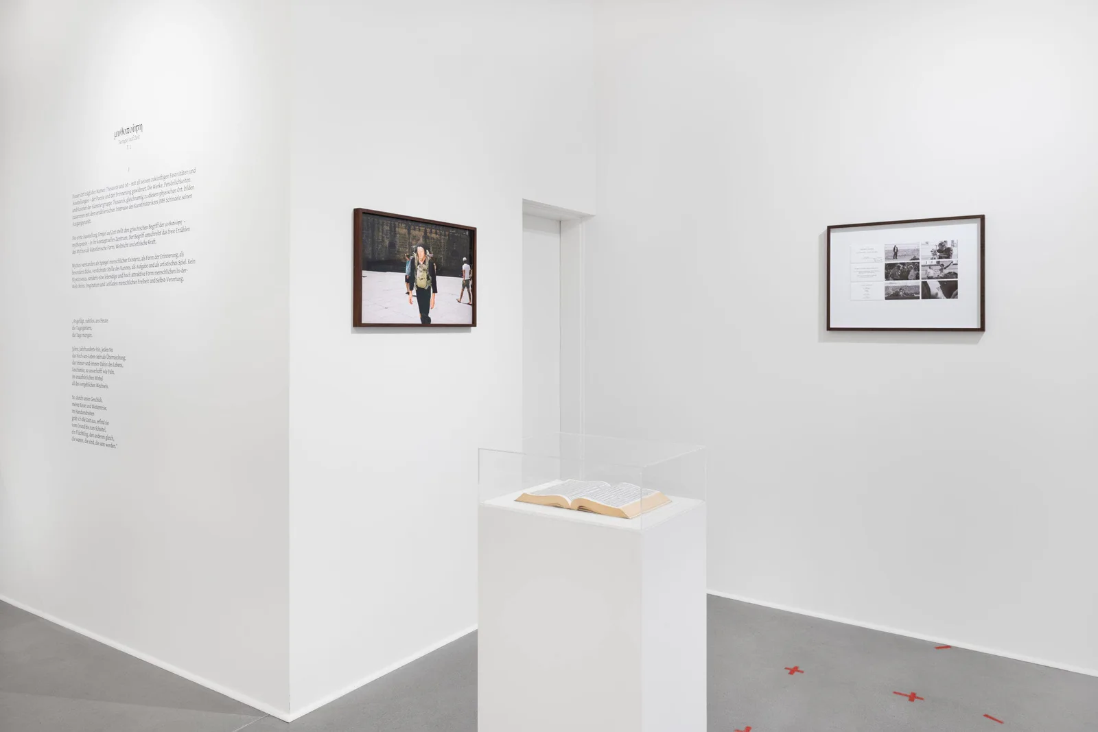 Ausstellungsansicht: weißer Raum mit Wandtext, zwei gerahmten Fotografien und einem flachen Sockel