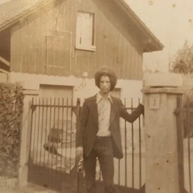 El Arbi Bouqdib in der Schweiz, frühe 70er: ein junger Mann im Anzug an einem Gartentor vor einem Holzhaus, Sepia-Foto