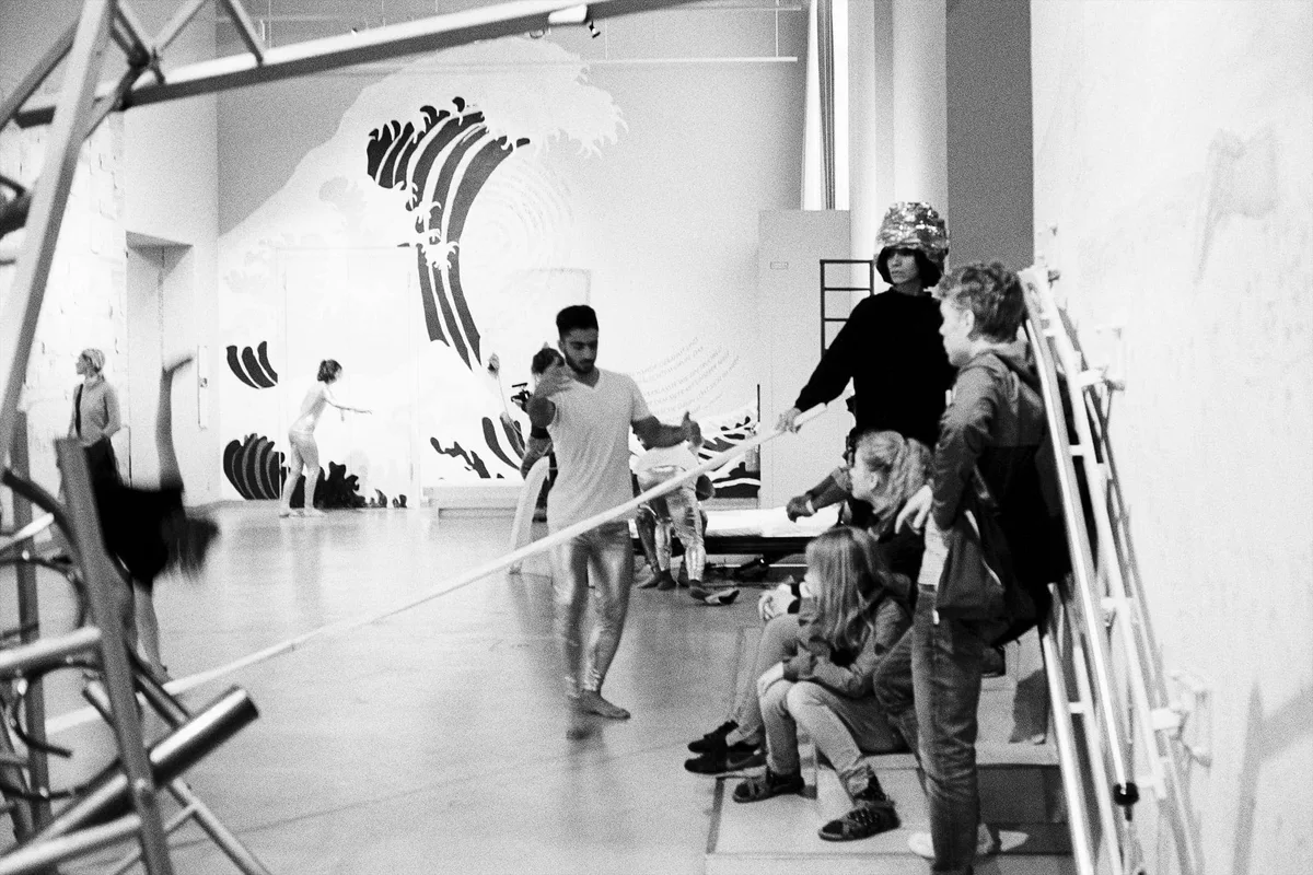 Schwarz-Weiß-Foto der Performance „MNEMOSYNE Prelude“: Besucher und eine Darstellerin zwischen Gerüststangen in einer Halle, an der Wand eine gemalte Welle