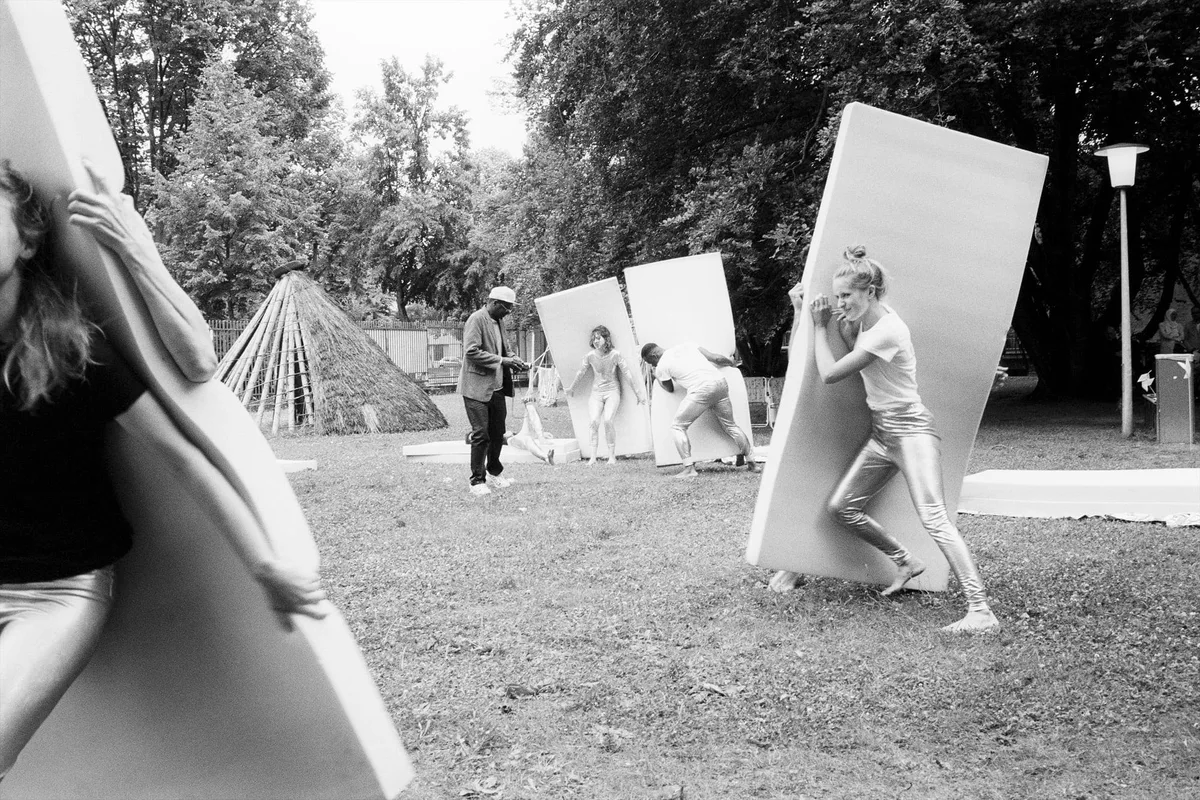 Performer tragen große weiße Tafeln über eine Wiese im Park, im Hintergrund ein Zeltgerüst