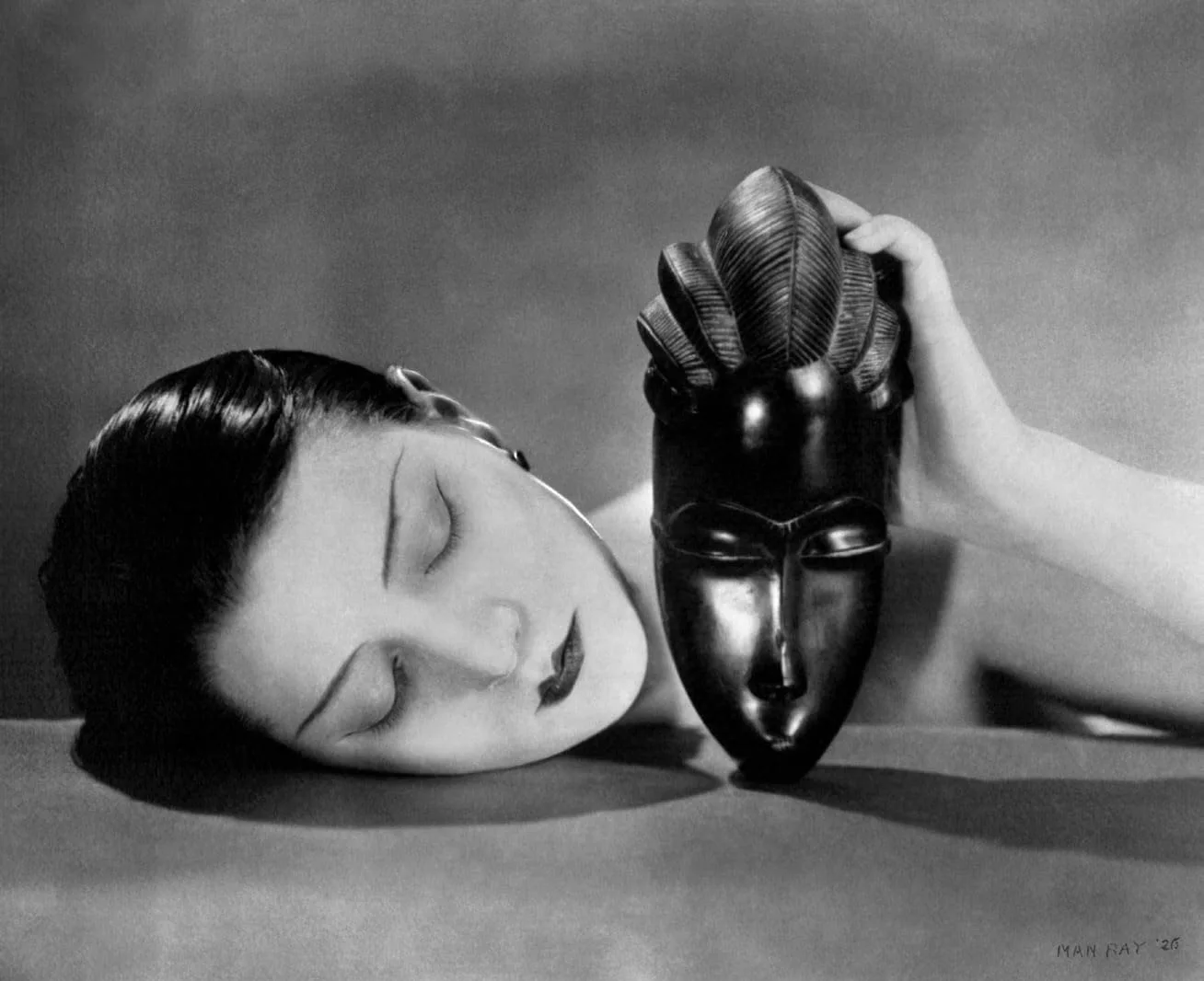 Man Ray, Noire et Blanche, 1926: Schwarz-Weiß-Fotografie einer Frau, die den Kopf auf einen Tisch legt und eine afrikanische Maske hält
