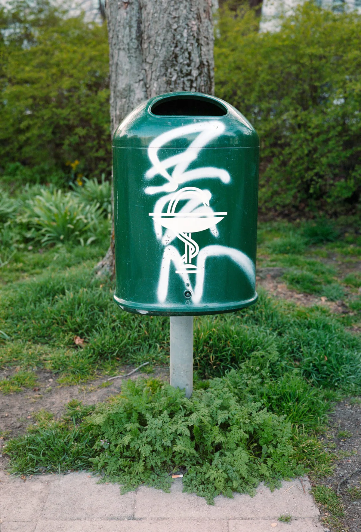 GABRIEL: grüner Abfalleimer mit Äskulapstab und Graffiti vor Gebüsch
