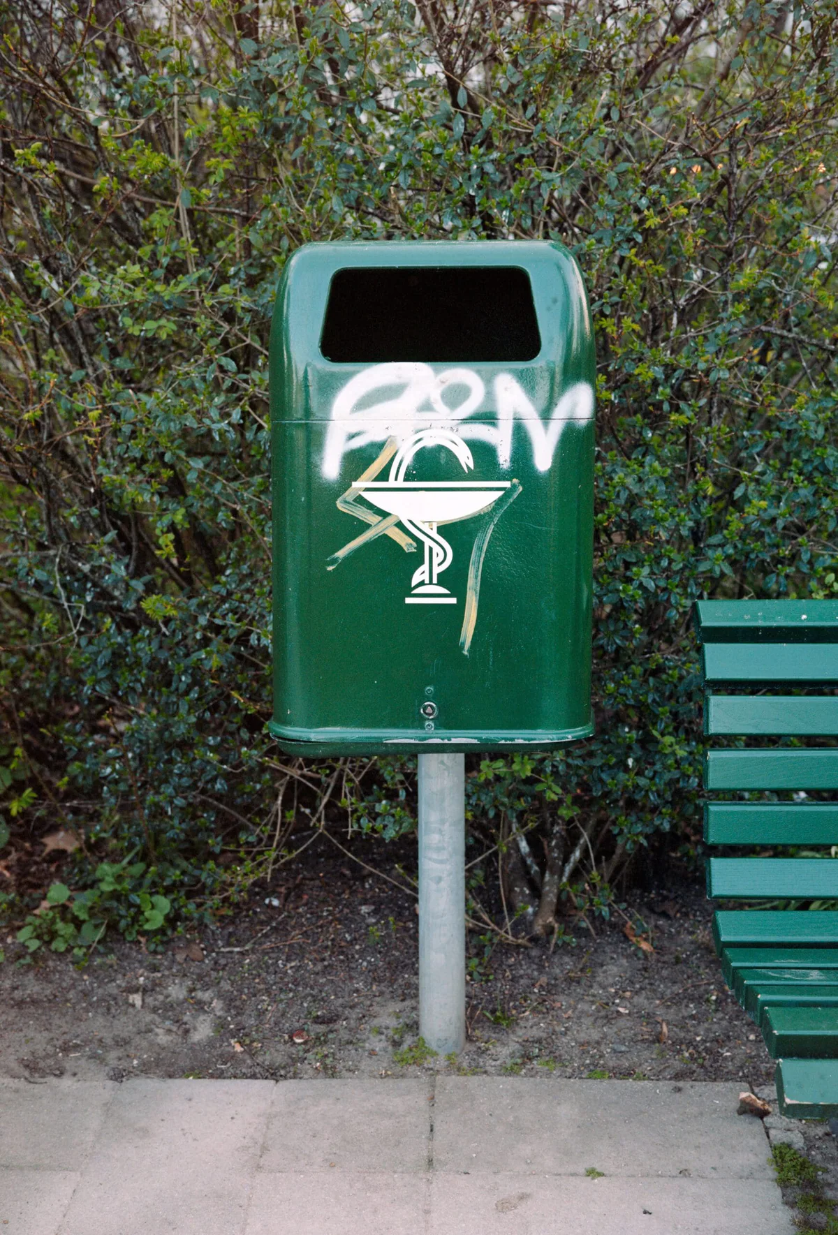GABRIEL: grüner Abfalleimer mit Äskulapstab und Graffiti neben einer Parkbank