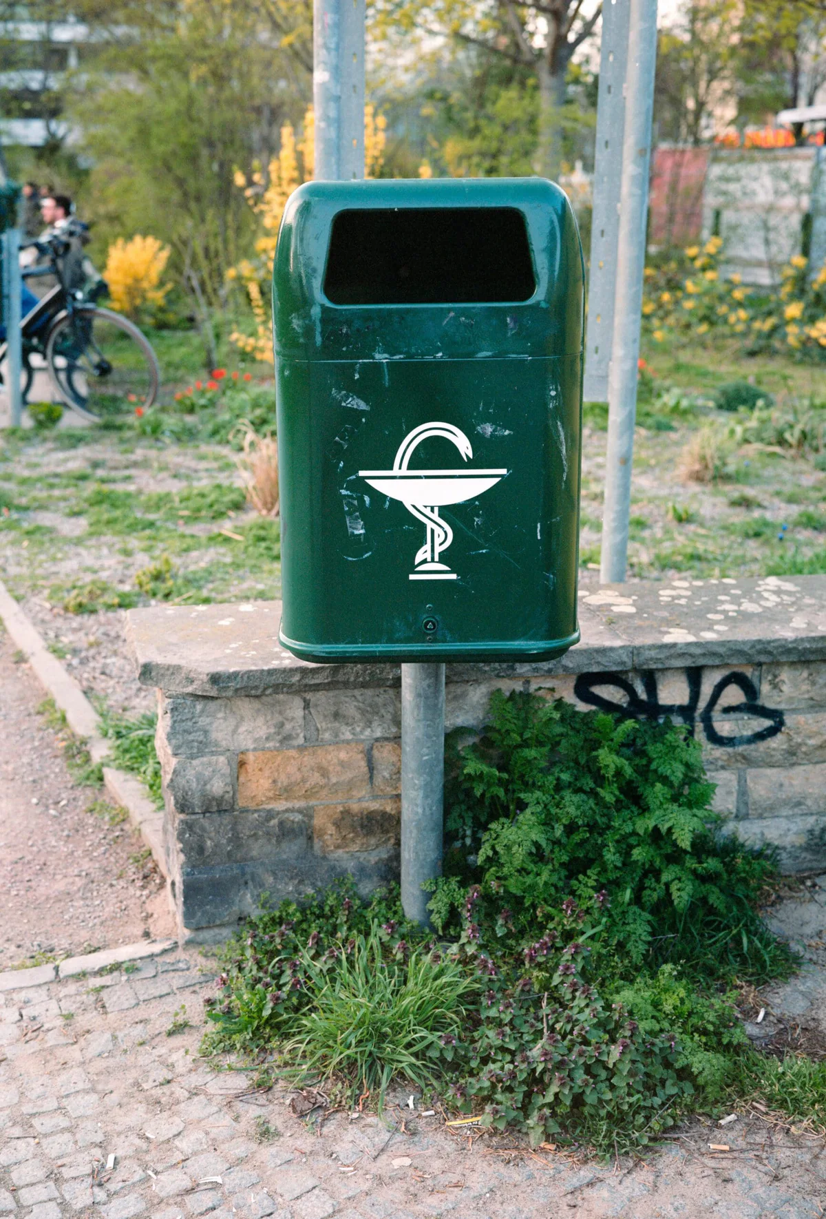 GABRIEL: grüner Abfalleimer mit Hygieiaschale vor einer Mauer mit Graffiti