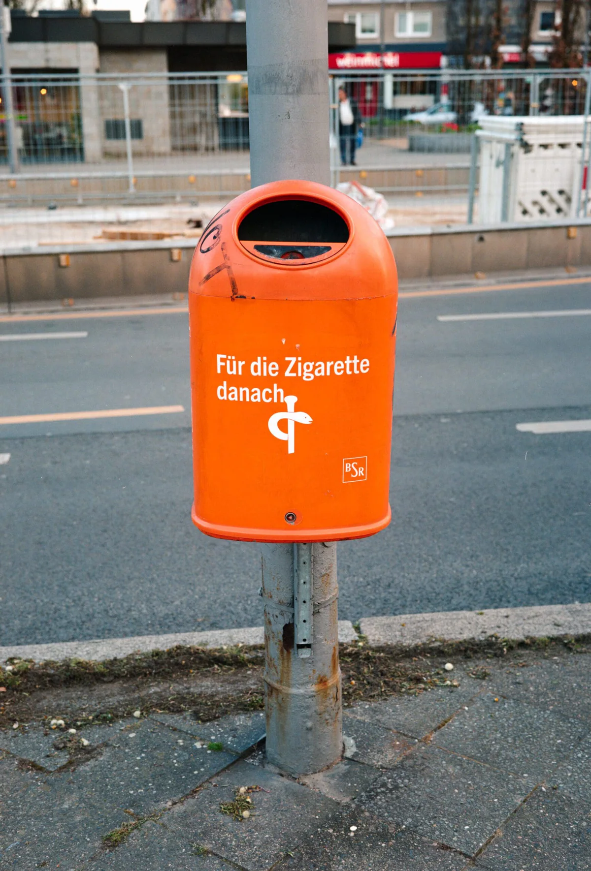 GABRIEL: oranger Berliner Abfalleimer mit Äskulapstab und der Aufschrift „Für die Zigarette danach“ an einer Straße