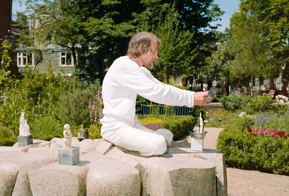 GABRIEL: Mann in Weiß sitzt mit kleinen Figuren auf einem Steinbrunnen in einem Garten