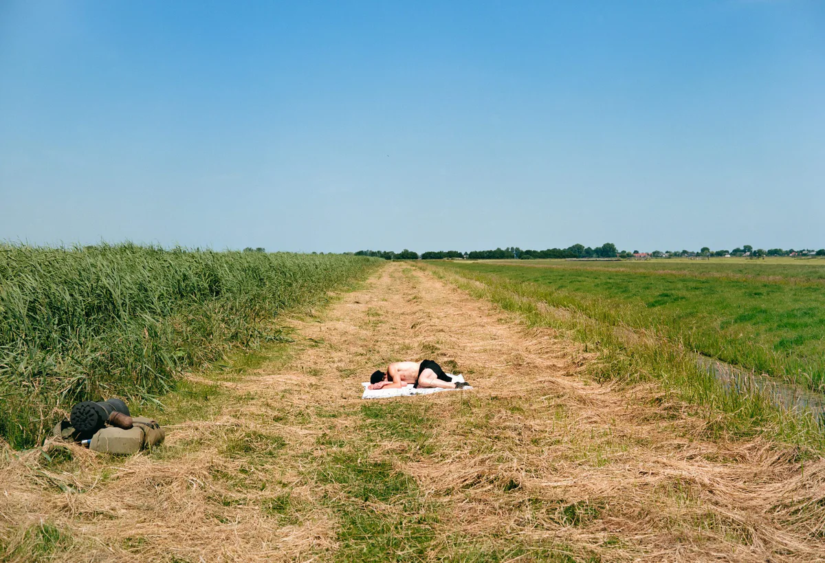 GABRIEL: Mann liegt auf einem weißen Tuch auf einem Feldweg zwischen Wiesen unter blauem Himmel