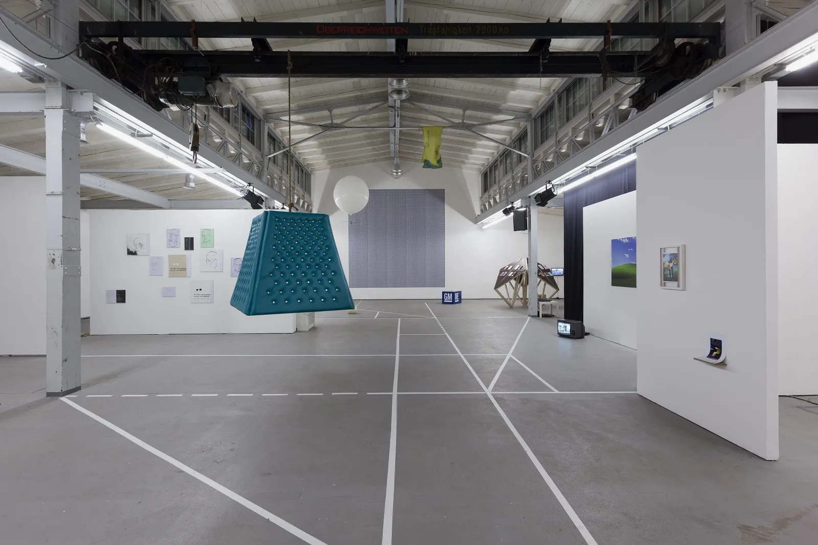 Installationsansicht: Industriehalle mit Bodenlinien, einem türkisfarbenen Pyramidenobjekt, Stellwänden und Bildern