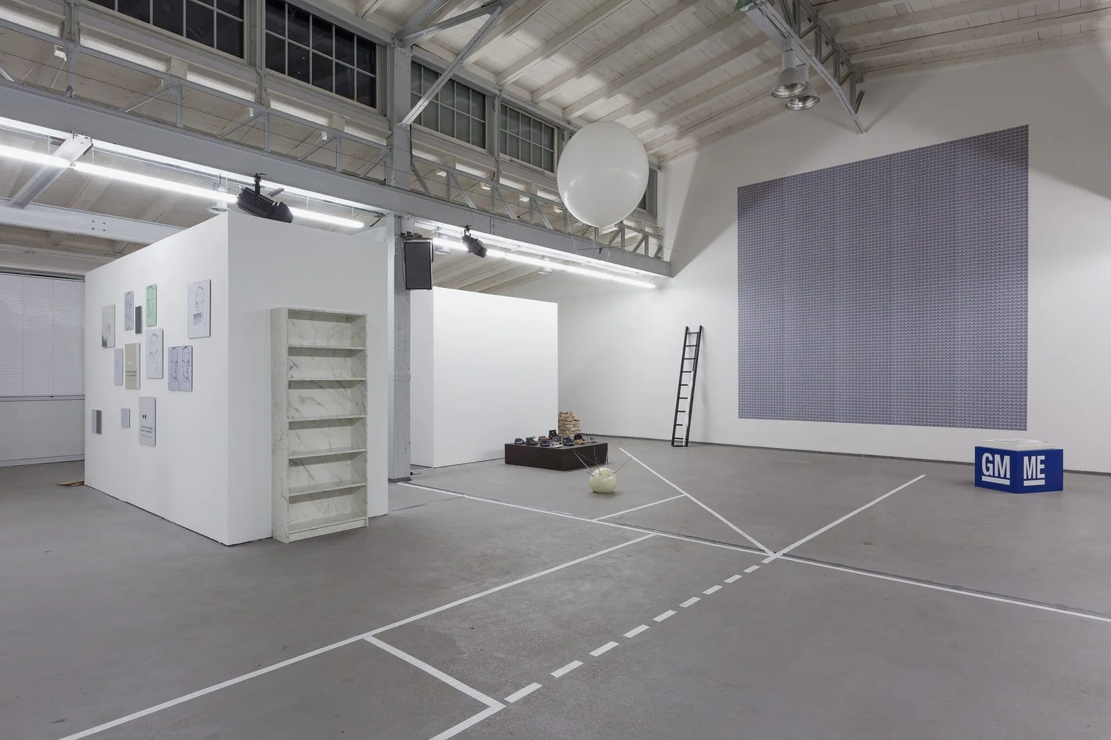 Installationsansicht: Industriehalle mit Bodenlinien, einer Leiter an einer grauen Wandfläche und Stellwänden mit Bildern