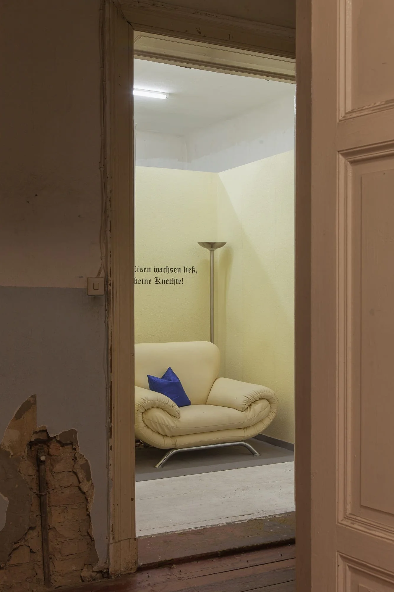 Installationsansicht: Blick durch eine Tür auf ein cremefarbenes Ledersofa unter einem Spruch an der Wand
