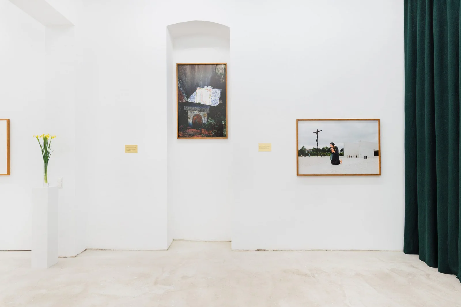 Marianische Antiphonen I – III, Ausstellungsansicht: weißer Raum mit gerahmten Fotografien, einem dunkelgrünen Vorhang und Blumen auf einem Sockel