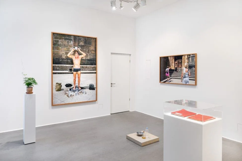 Installationsansicht: weißer Raum mit der Fotografie eines Mannes in Badehose, einer weiteren gerahmten Fotografie und einer Vitrine mit roten Büchern