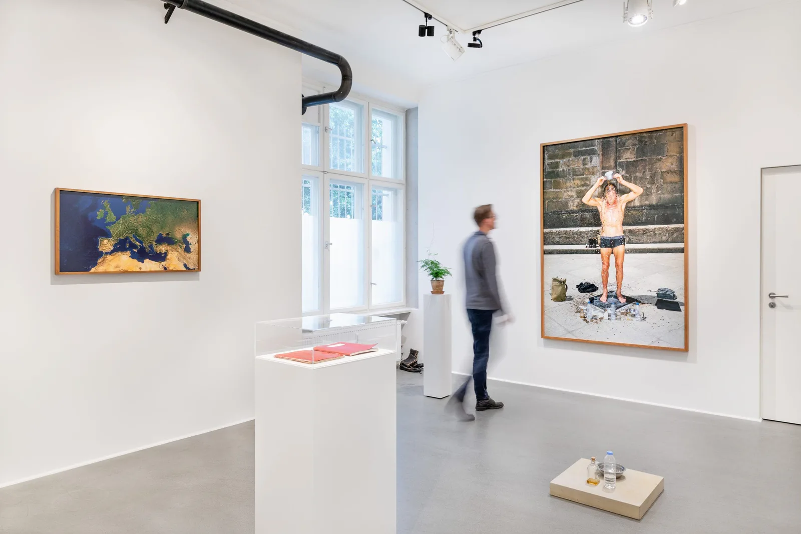 Installationsansicht: ein Besucher in einem weißen Raum mit Satellitenkarte Europas, Fotografien und einer Vitrine mit roten Büchern