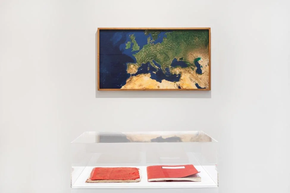 Installationsansicht: gerahmte Satellitenkarte Europas an der Wand, darunter eine Vitrine mit roten Büchern