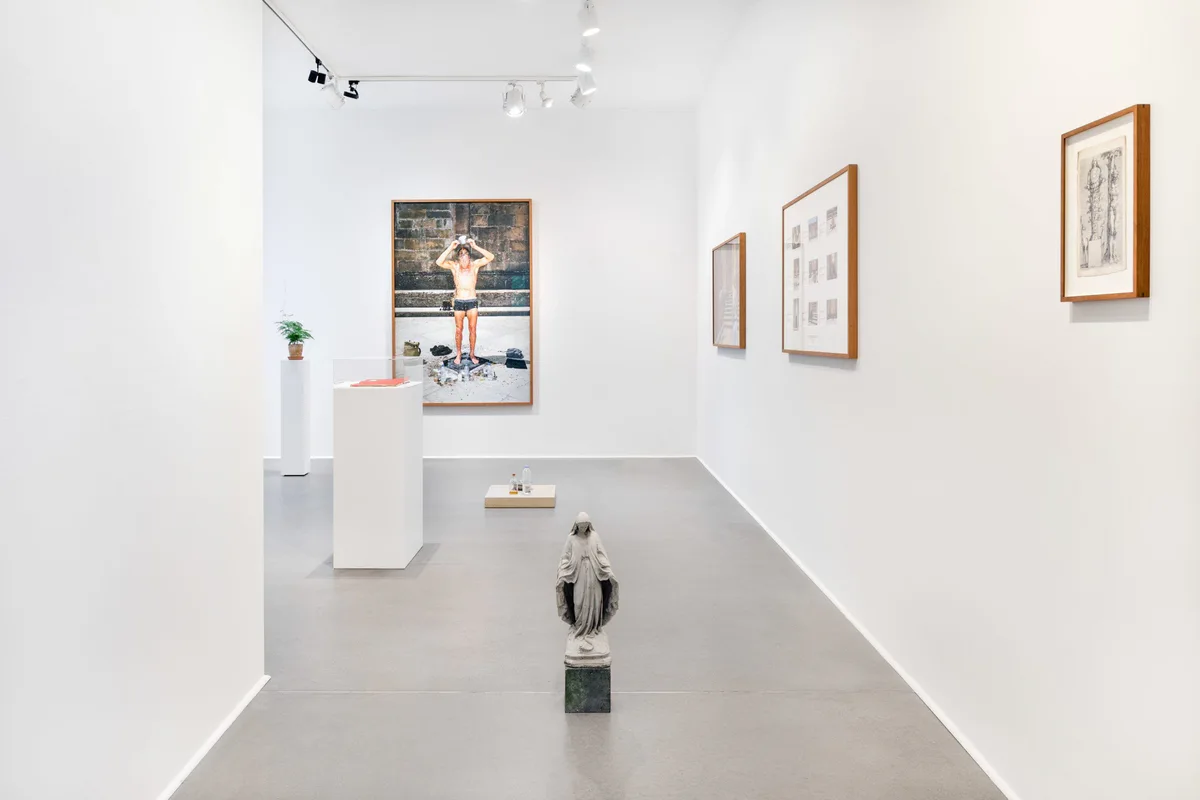 Marianische Antiphonen I, Ausstellungsansicht: Fotografie eines Mannes im Garten, kleine Blätter, ein Sockel mit Figur und ein Objekt am Boden