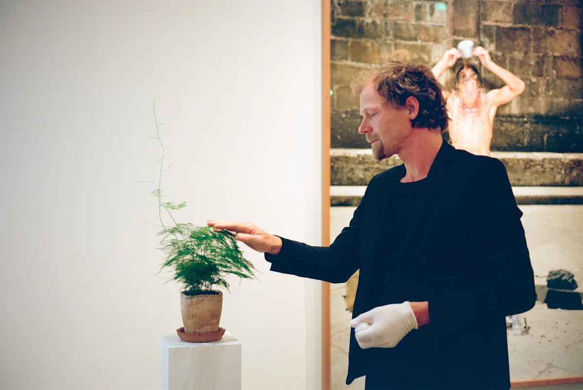 GABRIEL, Ausstellung bei Thesaurós: Mann mit weißem Handschuh berührt eine kleine Pflanze auf einem Sockel vor einer Fotografie