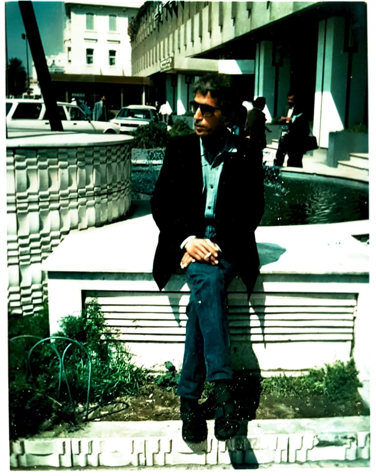El Arbi Bouqdib in a dark jacket by a fountain in a city, old colour photograph