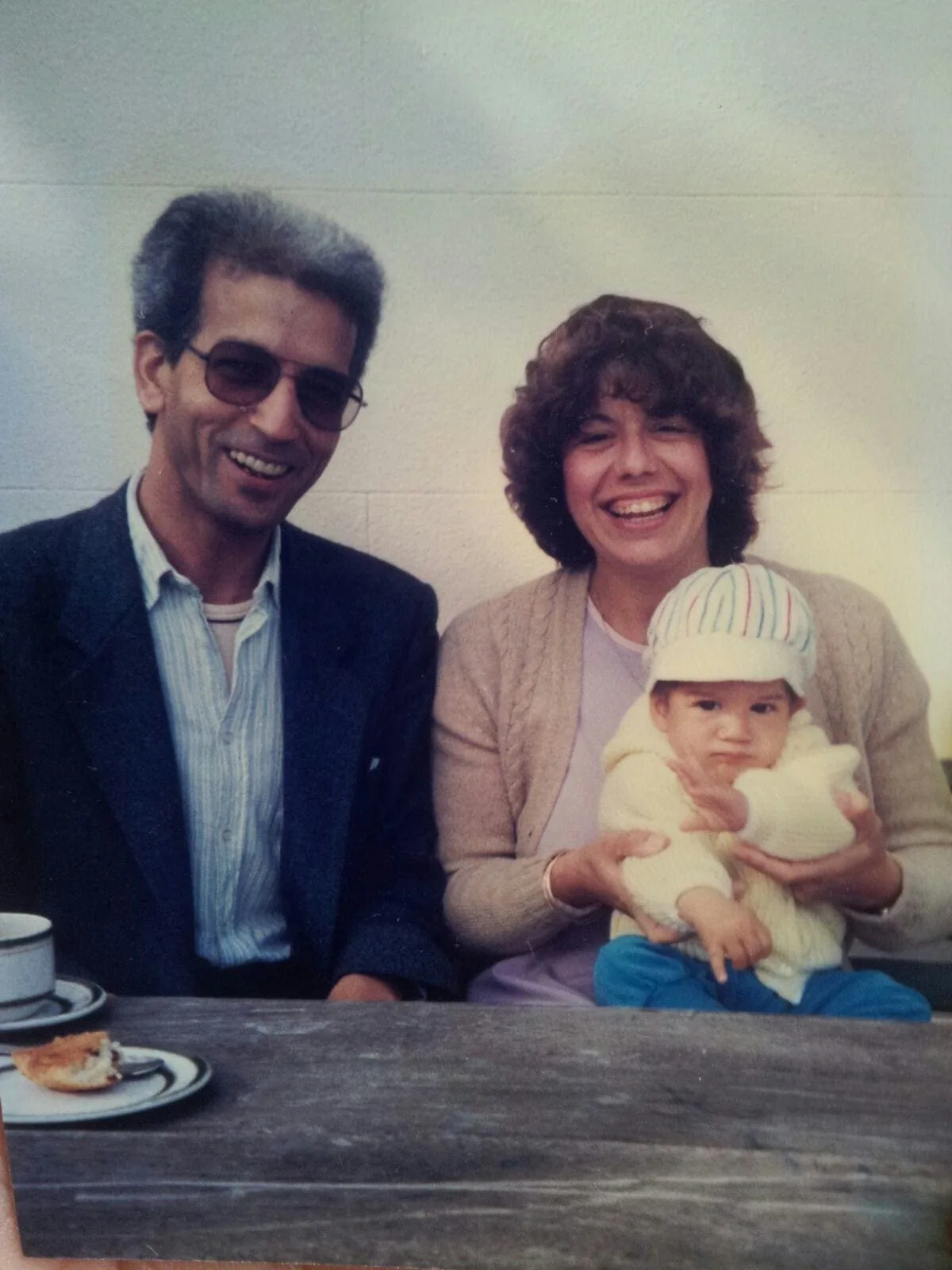 El Arbi Bouqdib in sunglasses beside a woman holding a baby at a café table, old colour photograph